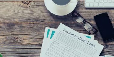 Insurance claim form. The form is on an old wooden table with a mobile phone, coffee, glasses and a keyboard. There is copy space to the right. Accident and insurance concept